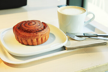 A fresh cinnamon roll and a cappuccino served on a white tray in a bright, airy cafe. This classic breakfast or coffee break is ready to be enjoyed.