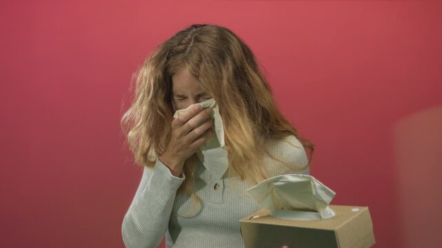 Woman lifting tissue experiencing sneezing in a sterile indoor environment beside a box with a blonde young sick sad cold expression.