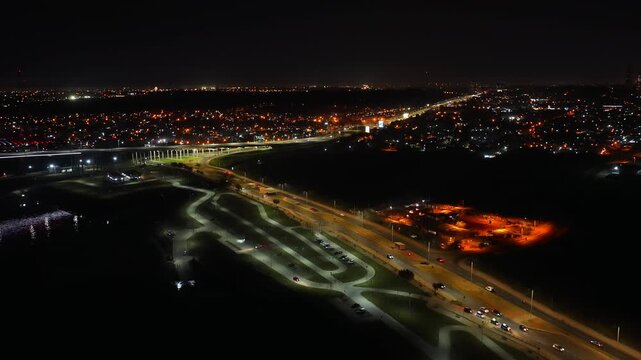 Drone video showcases Asuncion's Costanera and Heroes of Chaco Bridge. Night skyline glows with city lights below. Captures expansive urban landscape from above.