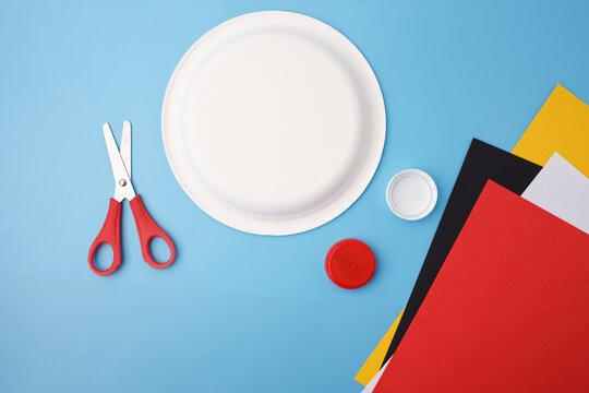 Paper plate, colored cardboard sheets, plastic bottle caps, and red scissors arranged on blue background. Flat lay composition showing DIY craft materials. Art and school project preparation