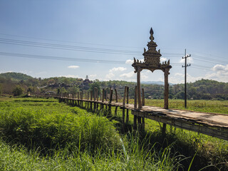 Panoramic view of the iconic Su Tong Pae Bridge, a traditional bamboo walkway stretching across vibrant rice fields towards a distant temple complex in Mae Hong Son, Thailand. A serene rural landscape