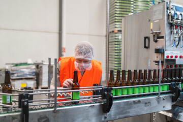 Brewery worker checking amber beer bottles moving on a conveyor belt in a processing plant,...
