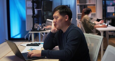 Focused Asian male programmer working late at night in modern tech office. Coding on laptop with data charts, developing software, analyzing code, or researching AI and cybersecurity solutions.