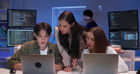 Focused group of young Asian developers working together on a coding project at night. Collaboration around laptops in a modern tech office with code screens and software development teamwork.