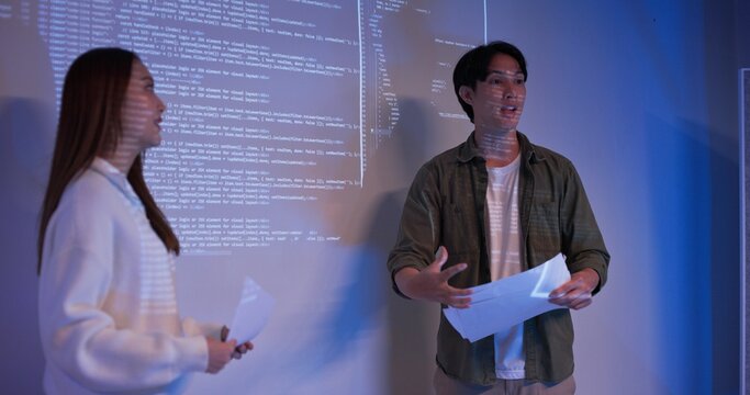 Young Asian software developers give team presentation in dark office environment. Explaining source code and project ideas under blue neon light. Modern IT teamwork and innovation in progress.