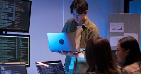 Group of young Asian developers collaborating during nighttime coding session. Team reviewing software issues and debugging source code in dark modern tech workspace. Innovation and teamwork in action