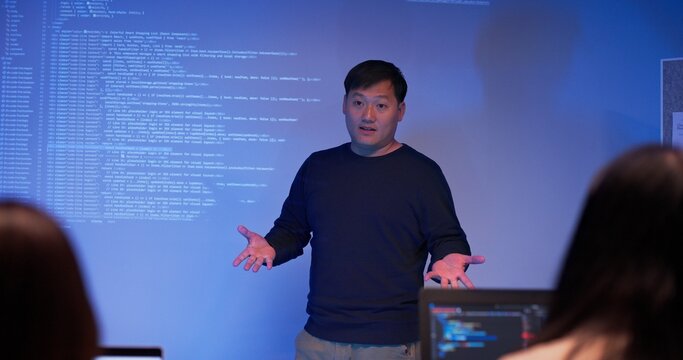 Asian male developer presenting source code to colleagues in a dark tech office. Team listening to coding explanation during software training, AI session, or cybersecurity presentation.