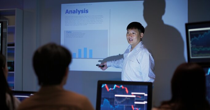 Confident Asian business professionals giving data-driven presentations to team in dark corporate office. Showing analytics charts, financial growth, and global strategy under projector light.