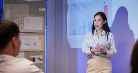 Professional Asian businesswomen lead data-driven presentations with global market insights in modern dark office. Team listens to financial reports, projections, and strategy under projector light.