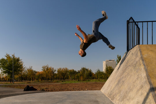 Male freerunner performs a backflip off a sloped concrete wall in an outdoor park, captured midair with extended limbs under clear skies, showcasing athletic precision and agility