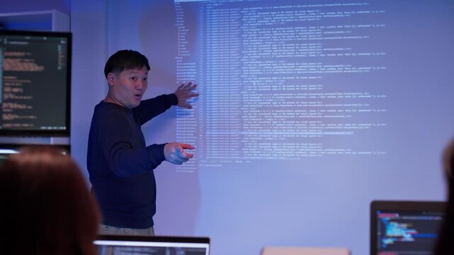 Asian male developer presenting source code to colleagues in a dark tech office. Team listening to coding explanation during software training, AI session, or cybersecurity presentation.