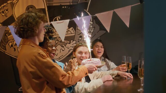 Young adult man with red curly hair clinking glasses with his friends and taking group selfie while celebrating birthday at bar