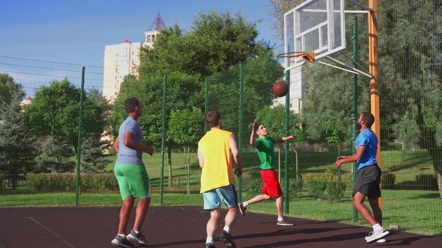 Active sporty fit basketball player making successful assist to African American male teammate, helping to score points with layup shot while multi ethnic sports teams playing game on outdoor court