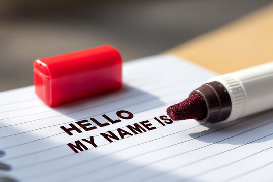A name tag is being written with a marker on lined paper, displaying the phrase "HELLO MY NAME IS" in bold letters with a red marker cap nearby