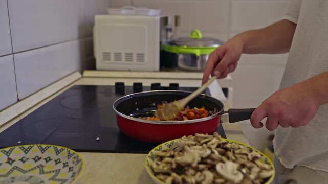 A person cooks chopped carrots and onions in a red pan on an induction cook top. Sliced mushrooms are ready to be added. This video includes the authentic sounds of cooking a healthy and delicious
