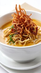 Creamy chicken Khao Soi in a white bowl, topped with crispy noodles and shallots, set against a warm beige background with gentle overhead lighting.
