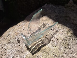 Broken clear glass pieces lying on a rough stone surface under bright sunlight. The sharp edges and...