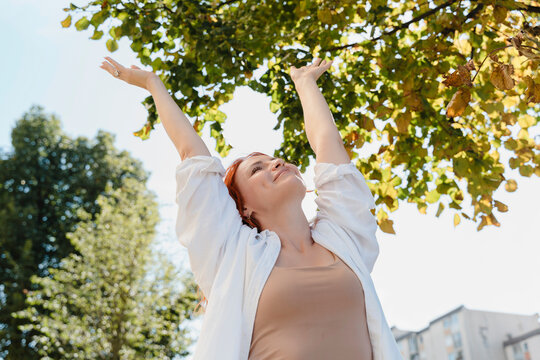 Red-haired woman with arms raised outdoors feeling optimistic and inspired - Powered by Adobe