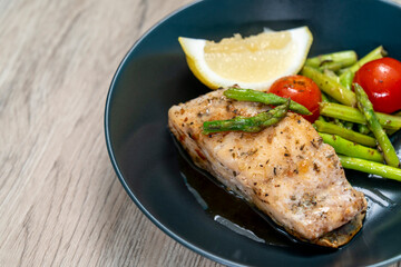 Red snapper steak, asparagus, tomatoes, and lemon on a black plate on wooden background. Table top view.