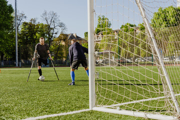 A man leans on crutches and is about to kick the ball while a man stands opposite him in front of...