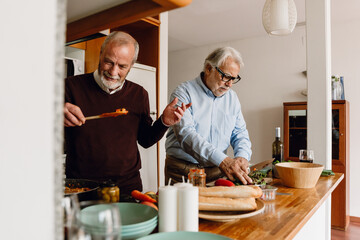 A man holds food on a spatula and smiles while a male friend stands next to him cutting vegetables