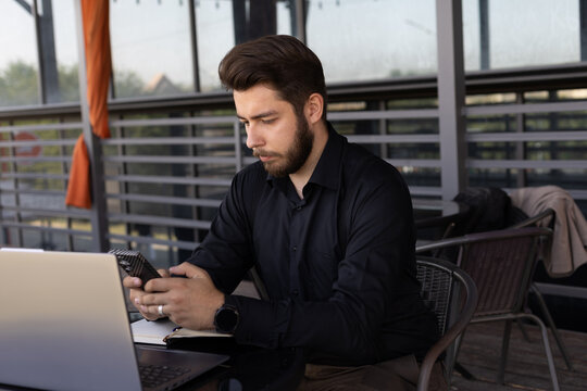 The man in the image is using his phone while sitting at a table with a laptop