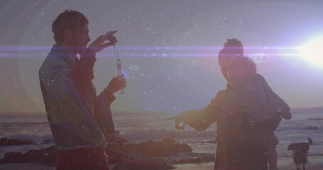Holding bubble wand, couple blowing bubbles on rocky shore at sunset, with lens flares and dog