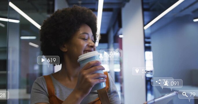 Woman in overalls holding coffee cup and tapping tablet in office, social media icons, copy space - Powered by Adobe