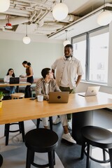 A male employee stands next to a male employee sitting at a desk while they talk