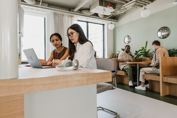 A female worker is talking and sitting at a table next to a female worker while they look at a laptop