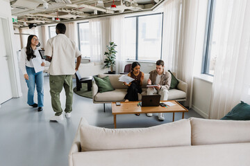 Fototapeta premium Male and female employees sitting on a couch holding a notebook and documents while male and female employees walking next to them talking