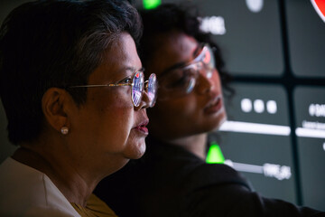 Two professionals collaborate beside a glowing digital dashboard, reflections on their glasses as they analyze data in a dark control room setting.