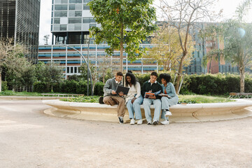 Obraz premium A group of four students sit and smile while looking in pairs at a laptop and notebook