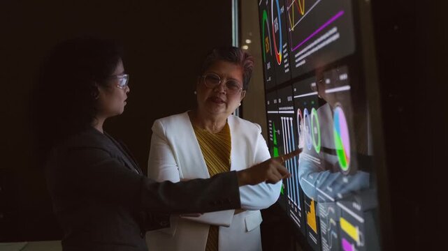 Two professionals review interactive data dashboards on a large touchscreen, discussing insights and strategy in a dark office with charts, graphs, and metrics. 4k video