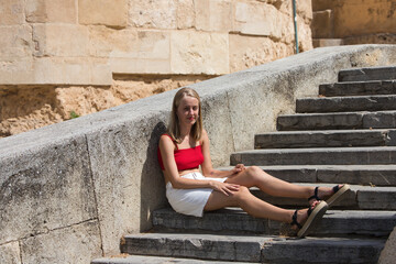 A young, beautiful blonde woman with make-up, blue eyes, a red top and a white skirt is sitting on the steps of a city park. The woman is on holiday in the city.