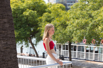 A young, beautiful blonde woman looks out from a viewpoint in seville to see the river that runs through the city. The woman is looking in different directions, enjoying the views.