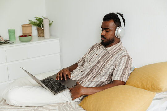 A man scrolls a laptop and listens in headphones while lying in bed
