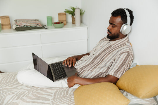 A man sits on a bed and listens in headphones while scrolling on a laptop