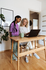 The daughter is standing next to the mother who is sitting at the table while they are looking at the laptop