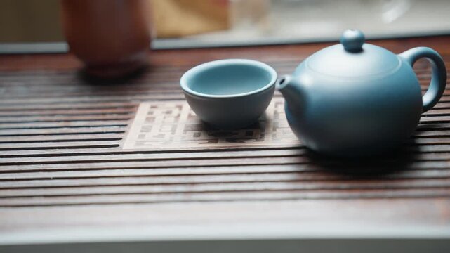 Slow motion video moving left to right: focus starts on a bamboo tea tray, then shifts to a blue clay teacup and finally a blue clay teapot, with blurred background.