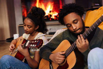 A man leans on a sofa and sits next to a woman while they play guitars