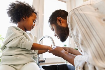 The father is standing and holding the hands of his daughter who is sitting on the table while they are washing their hands