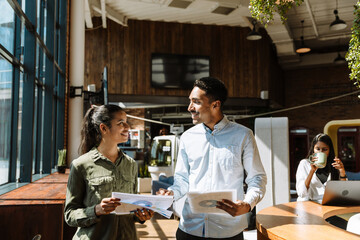 A male worker holds a folder while walking next to a female worker holding charts while they smile and look at each other