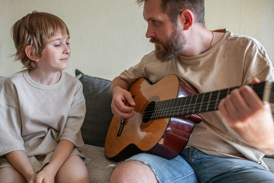 Bearded father and son sitting on sofa, man playing acoustic guitar while child watching with interest. Family bonding, father's day, music education, quality time at home concept. - Powered by Adobe