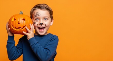 Happy young child boy with Halloween face paint and a carved pumpkin jack-o-lantern. Autumn holiday celebration and festive season tradition. Kid in studio on orange background with copy space
