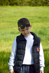 A boy standing against the backdrop of a green meadow on a sunny day. The child smiles slightly and looks away, creating a sense of naturalness and lightness