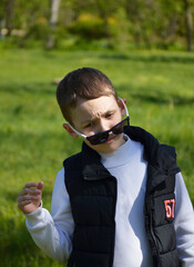 A boy standing against the backdrop of a green meadow on a sunny day. The child smiles slightly and looks away, creating a sense of naturalness and lightness