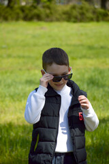 A boy standing against the backdrop of a green meadow on a sunny day. The child smiles slightly and looks away, creating a sense of naturalness and lightness