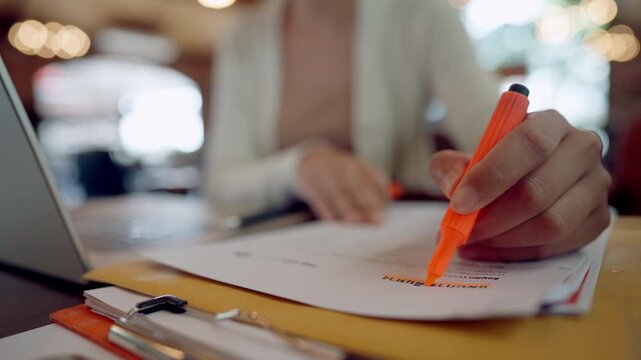 Close-up of woman highlighting business document with orange pen while working remotely with laptop.