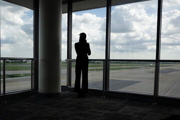 Fototapeta premium Silhouette traveler airport terminal window waiting departure runway clouds horizon. Silhouette traveler airport terminal window, waiting near departure gate with runway view dramatic clouds